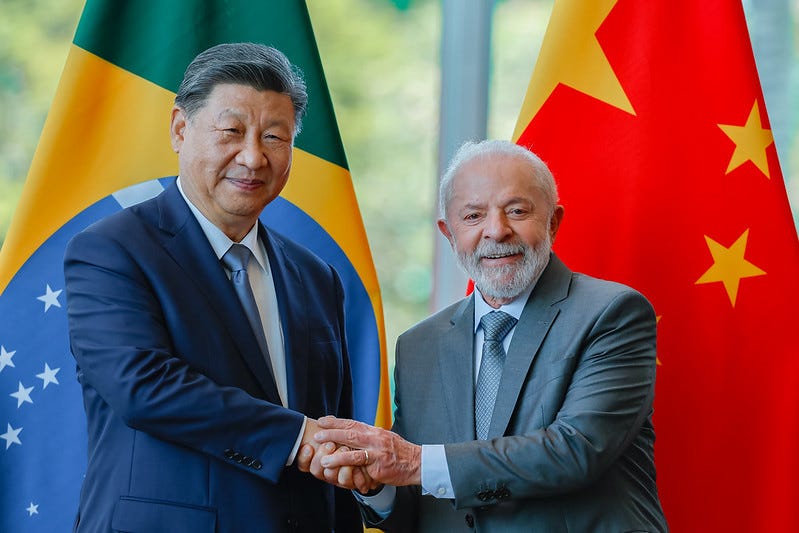 Fotografia em cores de Xi Jinping e Lula sorrindo e apertando mãos. Ao fundo, uma bandeira do Brasil (atrás de Xi) e um bandeira da China (atrás de Lula). A foto é de 2024.