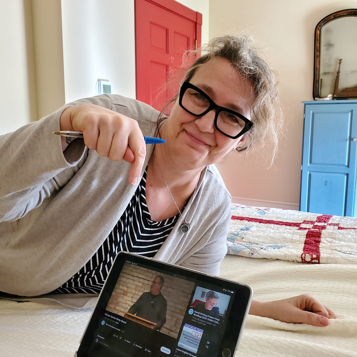 A white woman reclines on a bed, pointing to an iPad on which a white man stands at a microphone. 