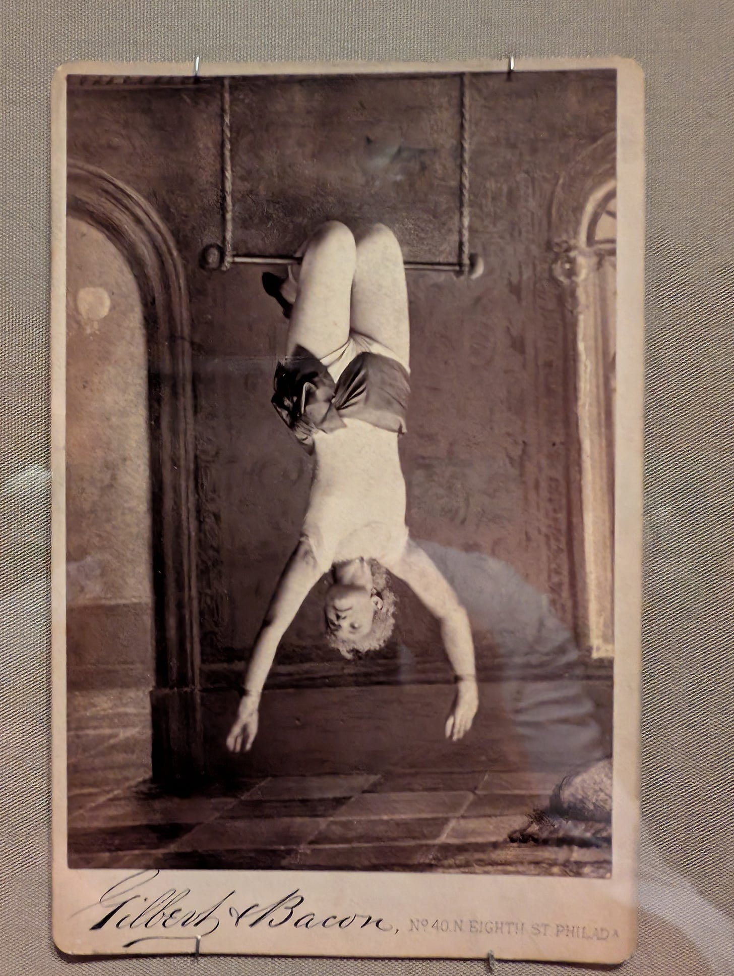 Sepia-toned photograph of Mlle. De Granville suspended upside down from a trapeze bar, arms extended, in a studio setting with painted architectural backdrop. Photograph by Gilbert & Bacon, 1880–86.