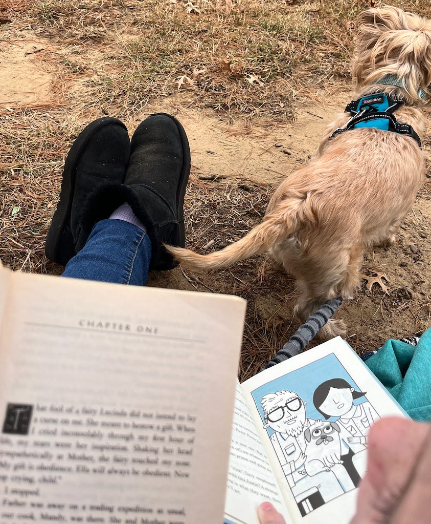 an adult and a child each reading a book next to a scruffy little dog on a blanket on the grass. 