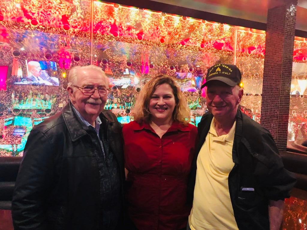 Jamie a blonde woman wearing a red blouse stands in the middle of two older men, the one on her right wearing a ballcap. They are posing in front of a colorful bar background at a restaurant. 