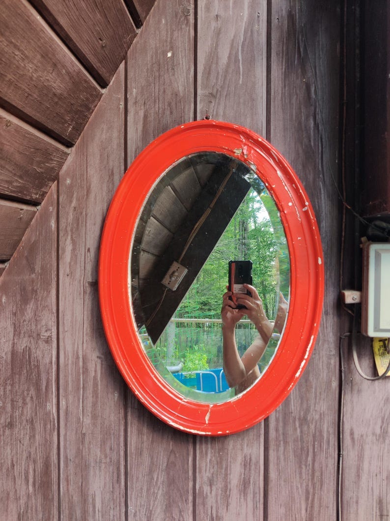 May include: An oval wall mirror featuring a bright orange painted metal frame with visible signs of wear and paint chipping. The reflective glass surface shows a distorted view of the surrounding environment, including trees and a building structure.