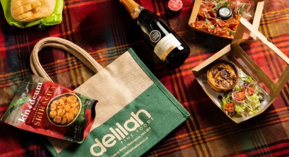 A hamper with a tote bag, a bottle of cremant, quiche, and bread, on a red checkered blanket
