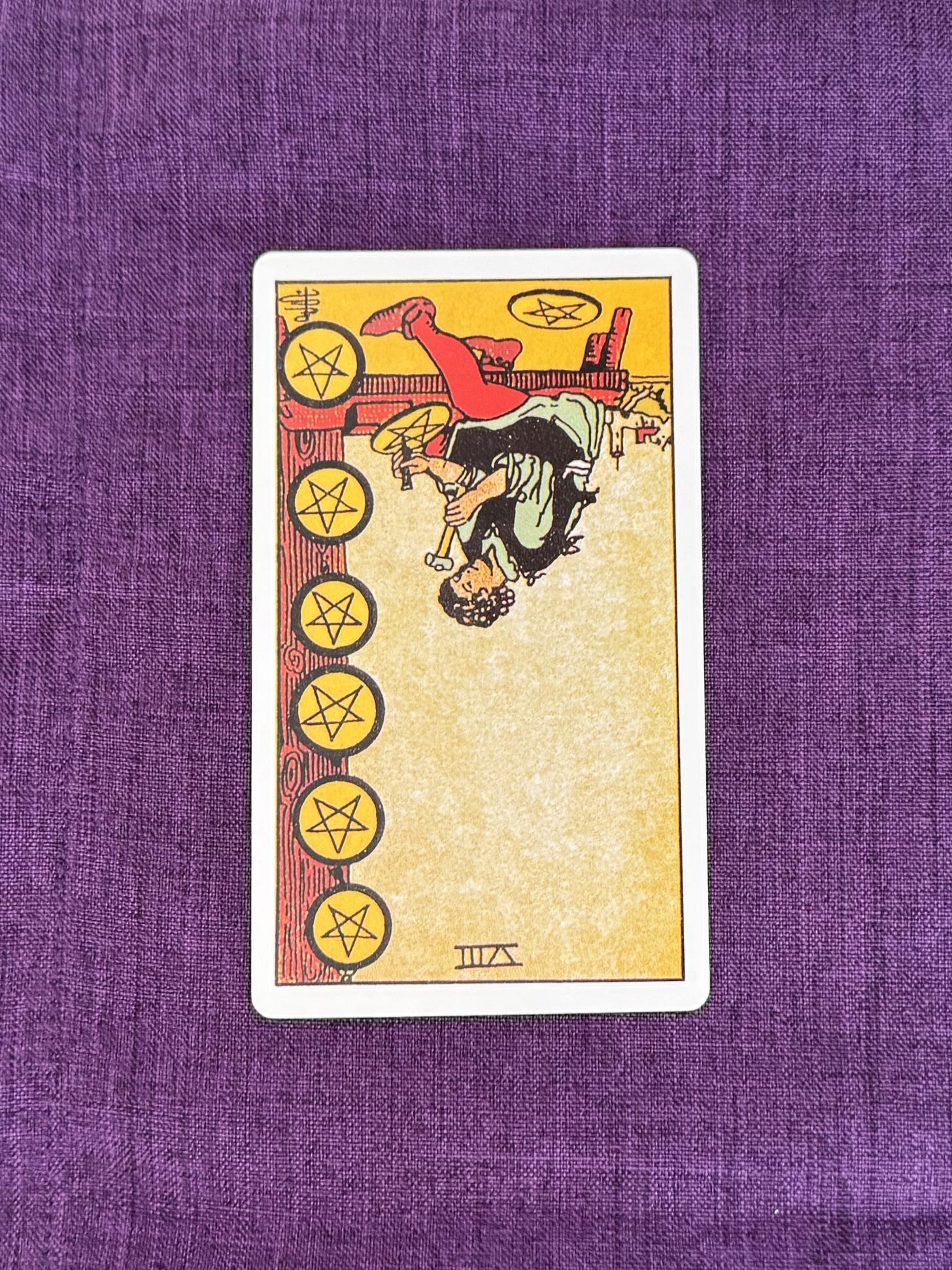 A figure sits on a wooden bench, working on a pentacle with a small hammer. Seven completed pentacles are displayed on the wall beside the figure, while the eighth is being worked on. The figure wears a simple tunic and apron. A wooden structure and a path are visible in the background beneath a clear sky. The reversed card means it's showing the image upside down.