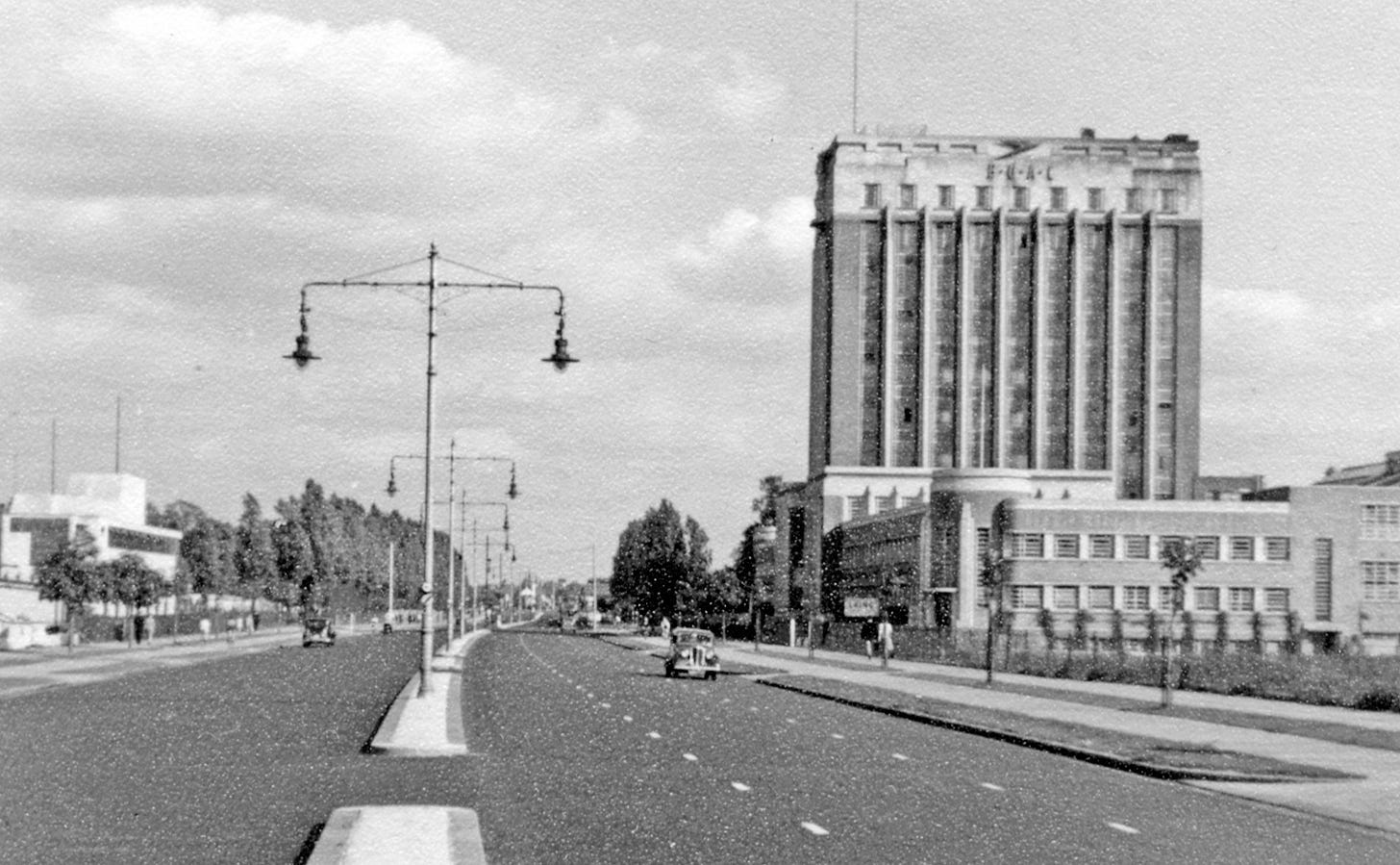 Black and white photograph of a wide dual carriageway passing between industrial buildings; an art deco office tower is on the right and a line of cast iron street lights run down the middle. A few vintage cars travel on the road. Black and white photograph of a wide dual carriageway passing between industrial buildings; an art deco office tower is on the right and a line of cast iron street lights run down the middle. A few vintage cars travel on the road.