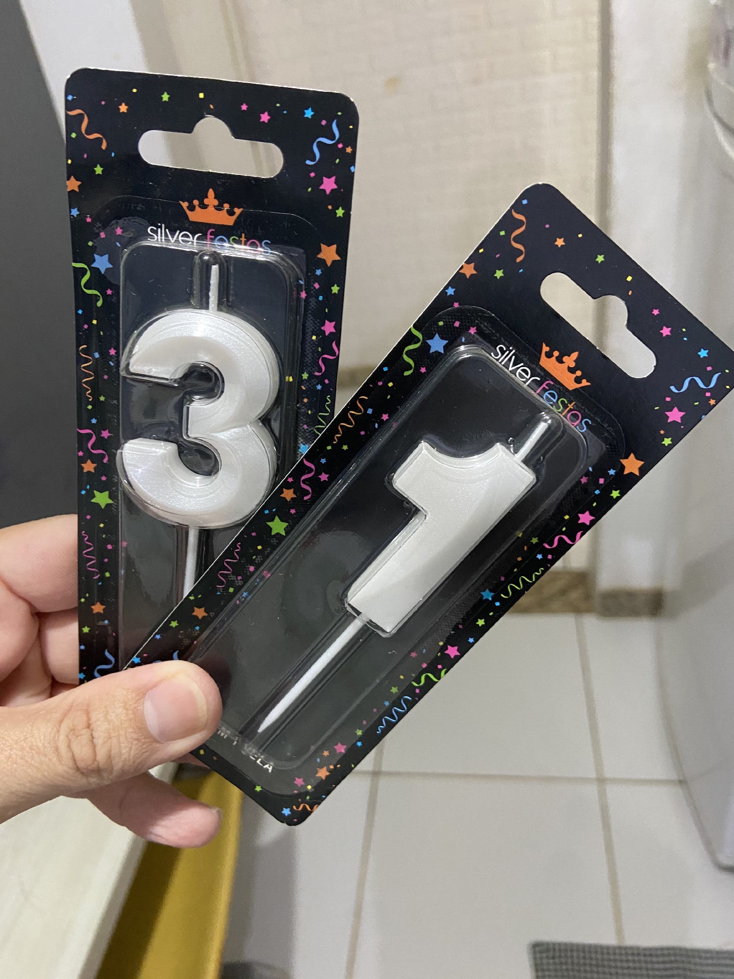 duas velas de aniversário em embalagens pretas com detalhea coloridos sendo seguradas entre o polegar e os demais dedos. uma vela é o "3" a outra é o "1"