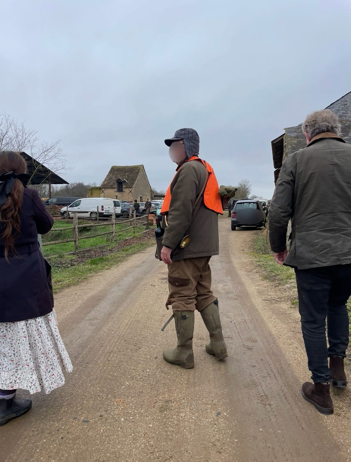 A man dressed for La Chasse in an orange vest and hat, a hunting horn under his arm and a bottle of wine tucked in his vest pocket, on a road in rural France.