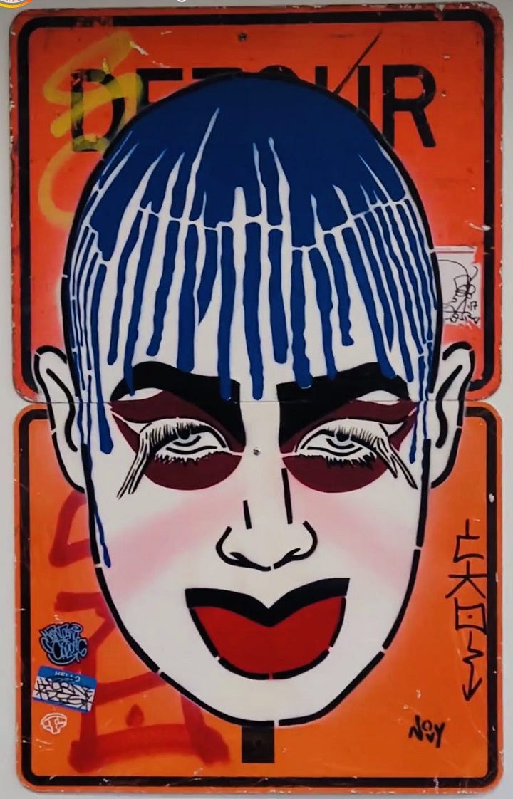 1) Street art on an orange "Detour" sign features a stylized face with blue paint for hair, bold red makeup, and exaggerated lashes, creating a vibrant and edgy vibe. This portrait is of Leigh Bowery, a famous queer person. There is more graffiti and tags behind Jeremy's work. 2) Street art on a dark black wall features a stylized portrait of a person with yellow hair and bold makeup. On the wall nearby, vibrant blue and purple graffiti tags. The portrait is of Divine, the drag queen. She has very high eyebrows, blue eyeshadow, and red lipstick, with a bright puffy yellow wig.