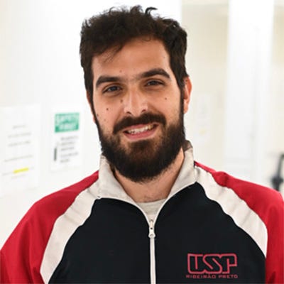 Homem de barba e cabelo escuro, vestindo um agasalho vermelho e preto com o logotipo “USP Ribeirão Preto” em um ambiente interno bem iluminado, como um laboratório ou corredor de instituição científica. Homem de barba e cabelo escuro, vestindo um agasalho vermelho e preto com o logotipo “USP Ribeirão Preto” em um ambiente interno bem iluminado, como um laboratório ou corredor de instituição científica.