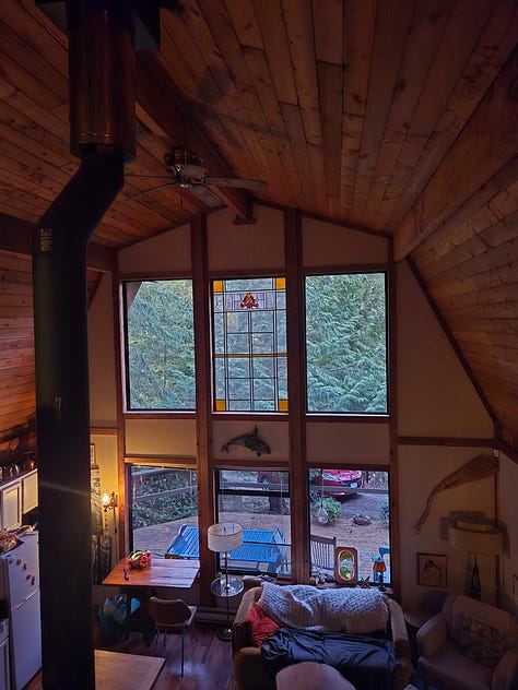 A laptop and a monitor on a desk with a manuscript shown on screen; a modest cabin with big windows in the woods surrounded by a deck; a view of the colourful rustic living space from the loft where I set up my desk. Two women sitting at a pub, and a desk with papers on it.
