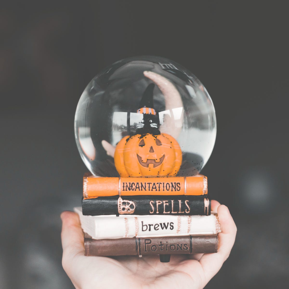 Hand holding a Halloween snow globe with a pumpkin and books labeled “incantations,” “spells,” “brews,” and “potions,” symbolizing the myths of secret formulas behind LinkedIn success.