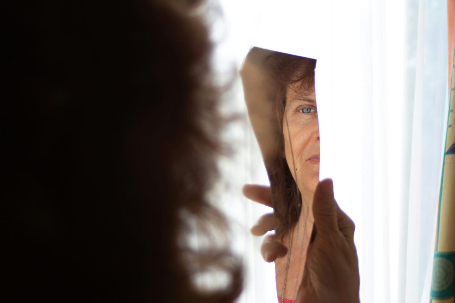 A mature woman, whose back is to the camera, holds a piece of broken mirror and is looking, unhappily, at half of her reflection.