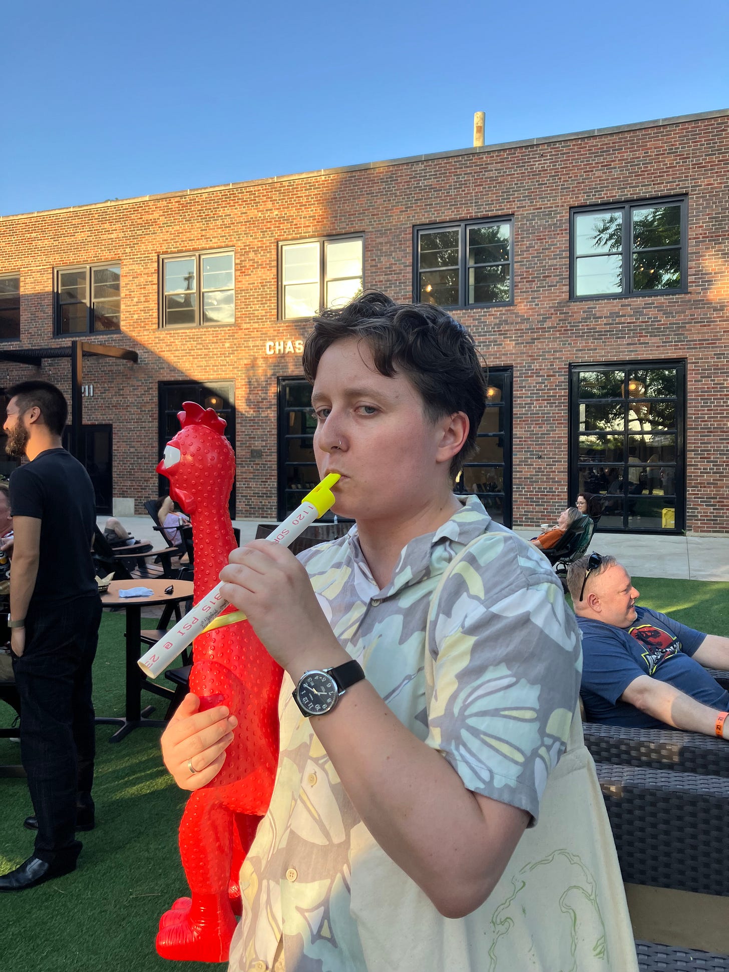 Hannah, looking unimpressed, toots a PVC pipe flute and holds a rubber chicken in a converted-industrial courtyard on a sunny day. Pianist Conrad Tao, in conversation with someone else and dressed in all black, is visible to the left; another man, middle-aged and in a T-shirt, lounges in a patio chair to the right.