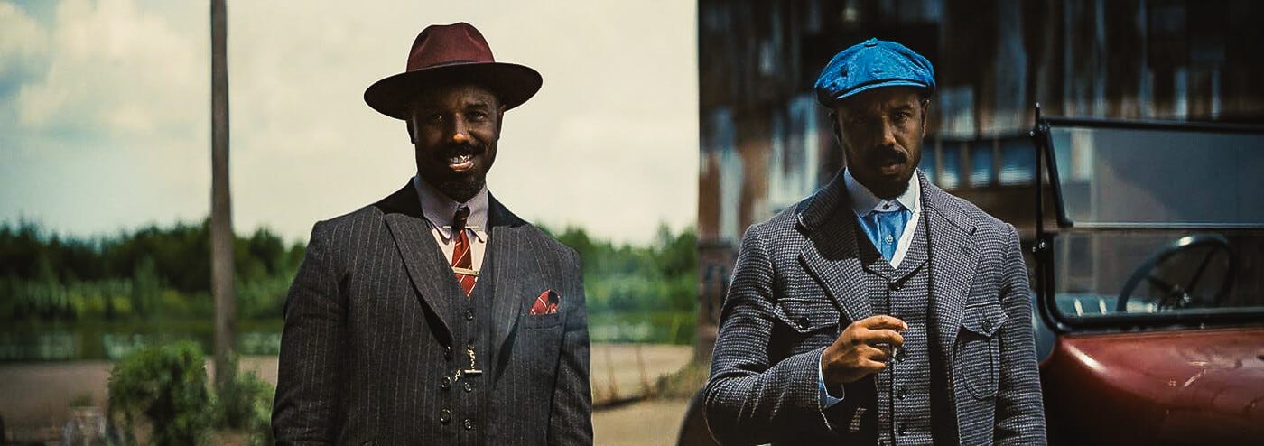 Michael B. Jordan as twin brothers in Sinners, dressed in vintage suits and standing beside a red convertible. One sports a red fedora and tie to match while smiling, the other in a blue jockey’s cap and matching shirt looking sternly ahead.
