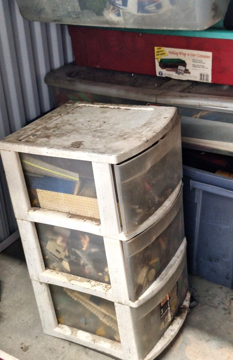 A dirt-covered three-drawer plastic storage unit filled with belongings sits among tubs and containers inside a crowded storage space.