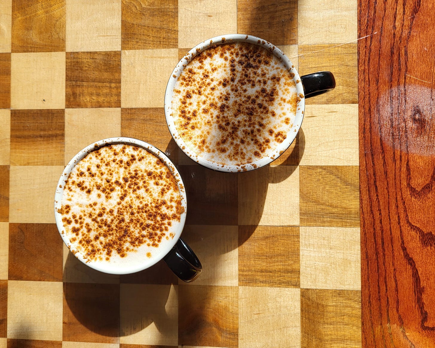 Two chai lattes in black mugs sit on a wooden table with a checkerboard pattern.