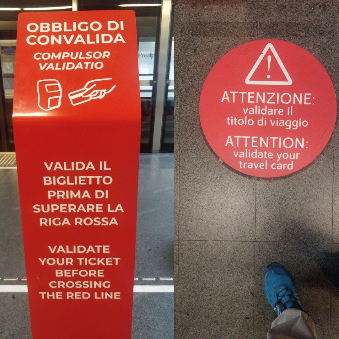 Due segnali di avviso rossi in una stazione della metropolitana o ferroviaria in Italia. Il cartello verticale a sinistra, con icone di un biglietto e di una mano, indica: "OBBLIGO DI CONVALIDA / COMPULSOR VALIDATIO" e "VALIDA IL BIGLIETTO PRIMA DI SUPERARE LA RIGA ROSSA / VALIDATE YOUR TICKET BEFORE CROSSING THE RED LINE". L'adesivo circolare sul pavimento a destra, con un punto esclamativo di avvertimento, ripete il messaggio: "ATTENZIONE: validare il titolo di viaggio / ATTENTION: validate your travel card". Si intravede un piede con una scarpa blu.