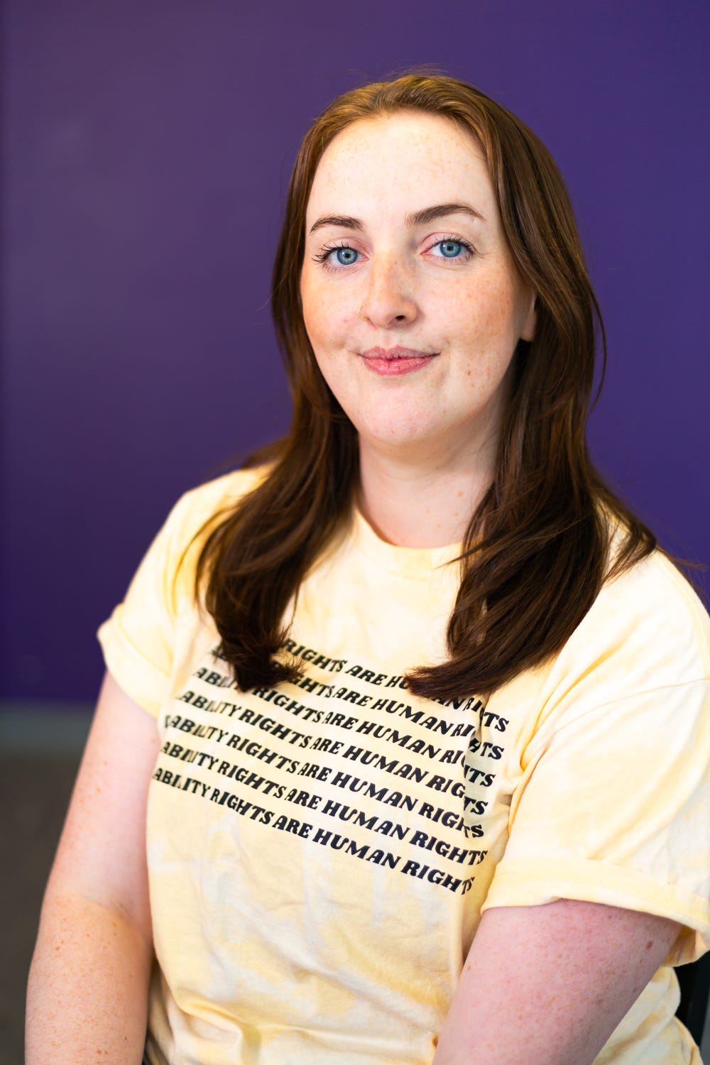 A portrait of Nicole, a young, white-skinned woman in her 20’s. She has auburn hair parted in the middle that goes past her shoulders and striking blue eyes with which she looks into the camera. She has the expression of a woman doing important advocacy work. She is wearing a yellow shirt that reads, “Disability Rights Are Human Rights”