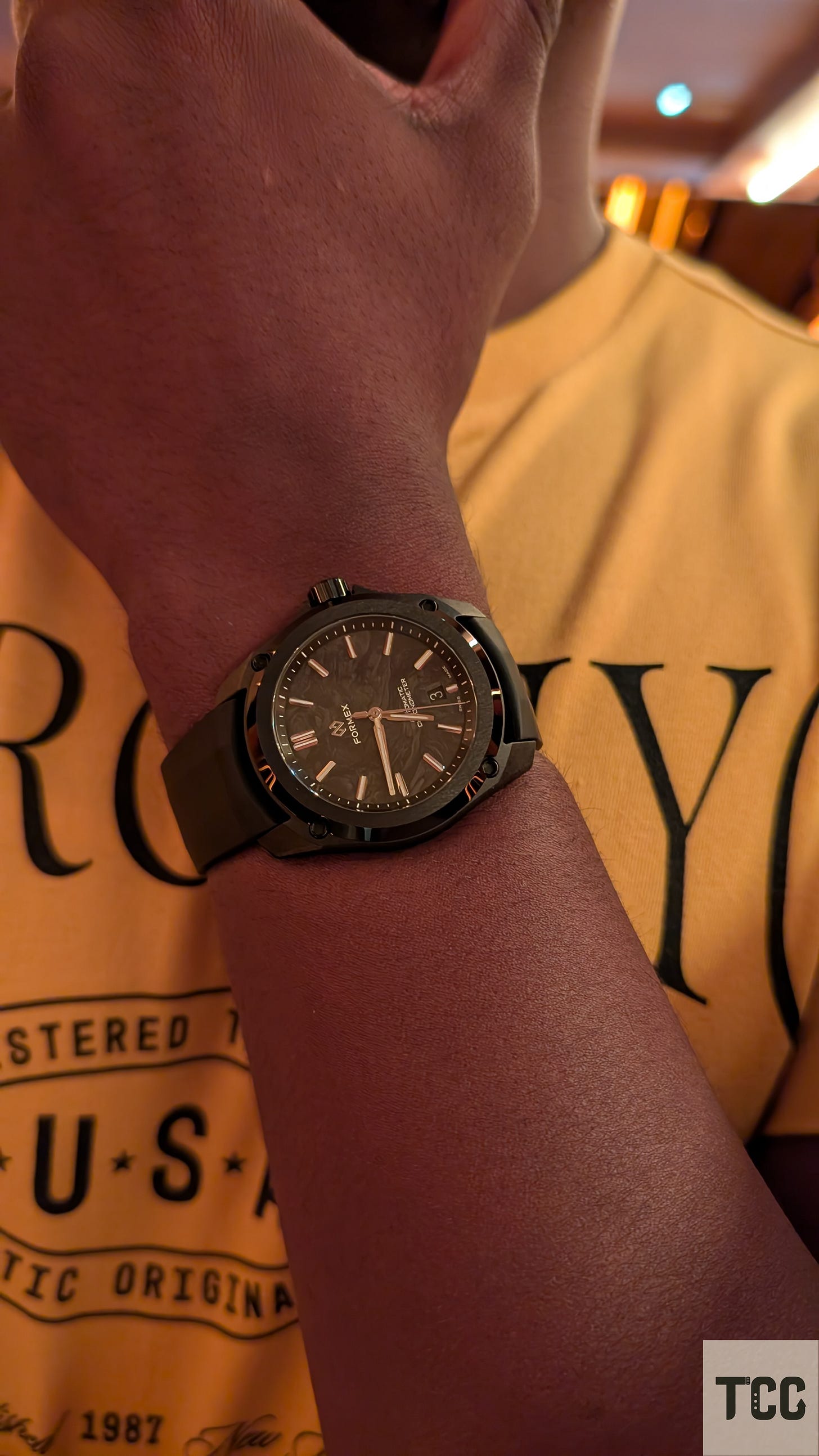 A close-up shot of a Formex Reef watch with a forged carbon dial on the wrist of a man wearing a yellow "AERO NYC" T-shirt. The watch is seen indoors under warm lighting, highlighting the intricate dial texture and bold case design.