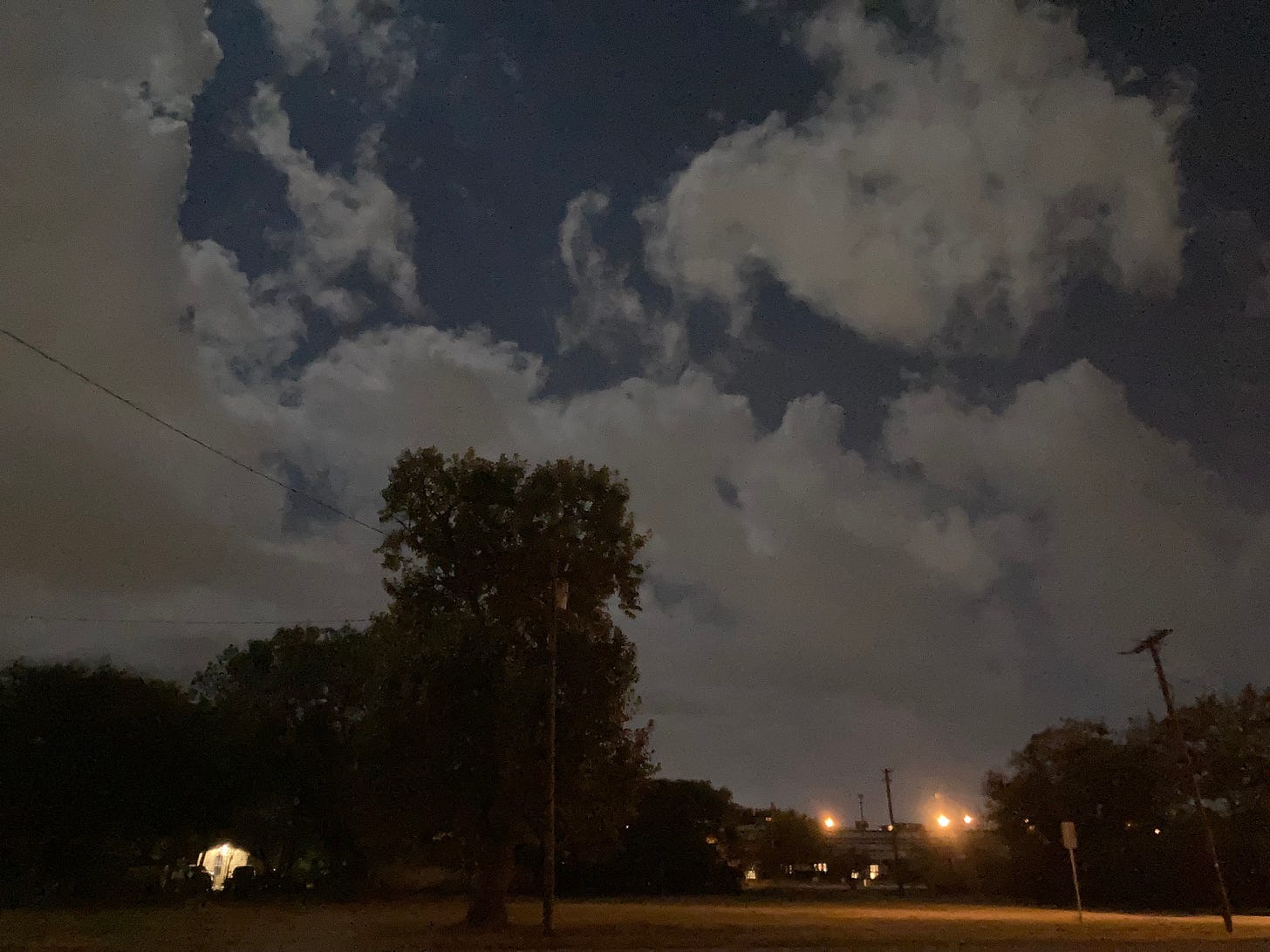 A few lights glare in the darkness shrouding the urban treeline, as clouds tower over the gloom.