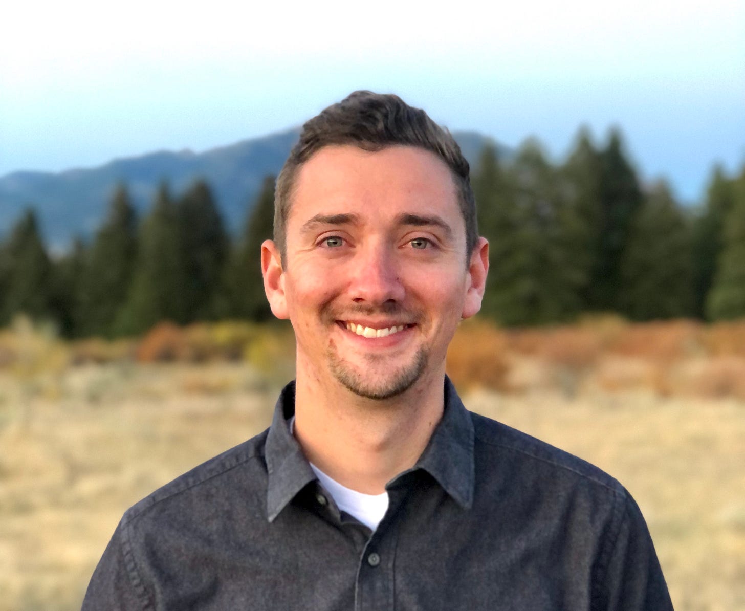 smiling man in dark gray button up at edge of woods