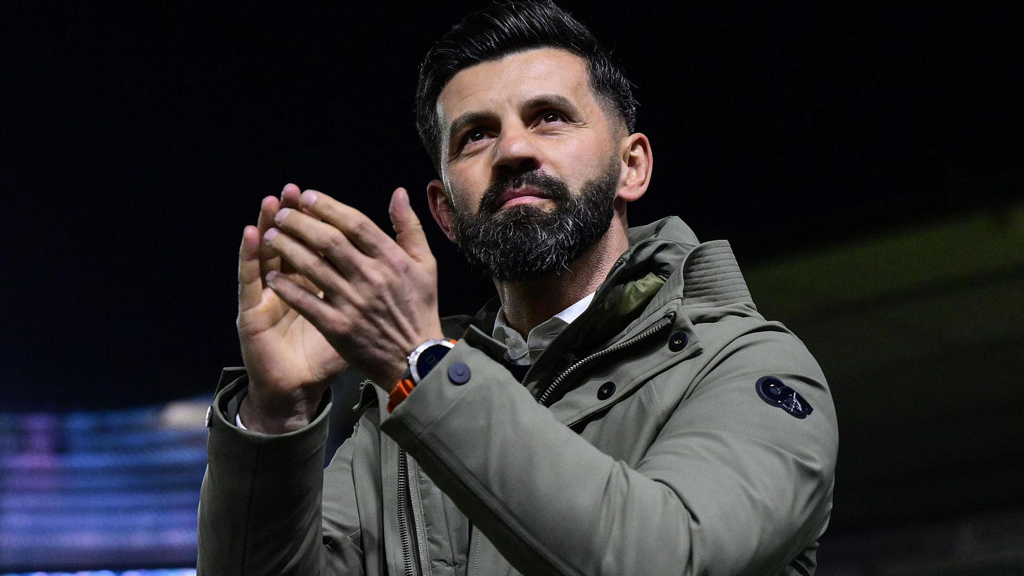 A close-up image of Miron Muslic looking up. He is wearing a grey jacket and is clapping his hands. A close-up image of Miron Muslic looking up. He is wearing a grey jacket and is clapping his hands.
