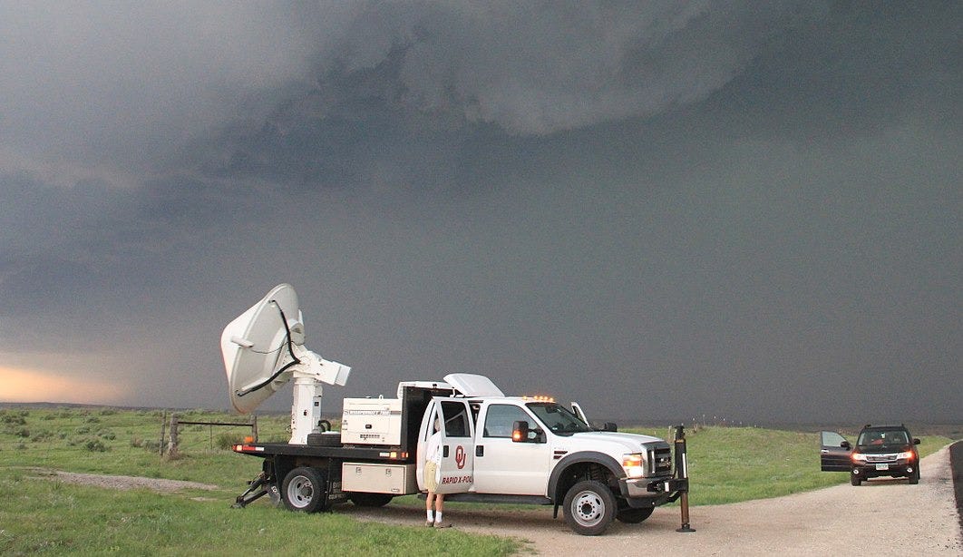 A white truck with a radar dish on its back parked next to a road in a field. In the back are very dark clouds. A white truck with a radar dish on its back parked next to a road in a field. In the back are very dark clouds.