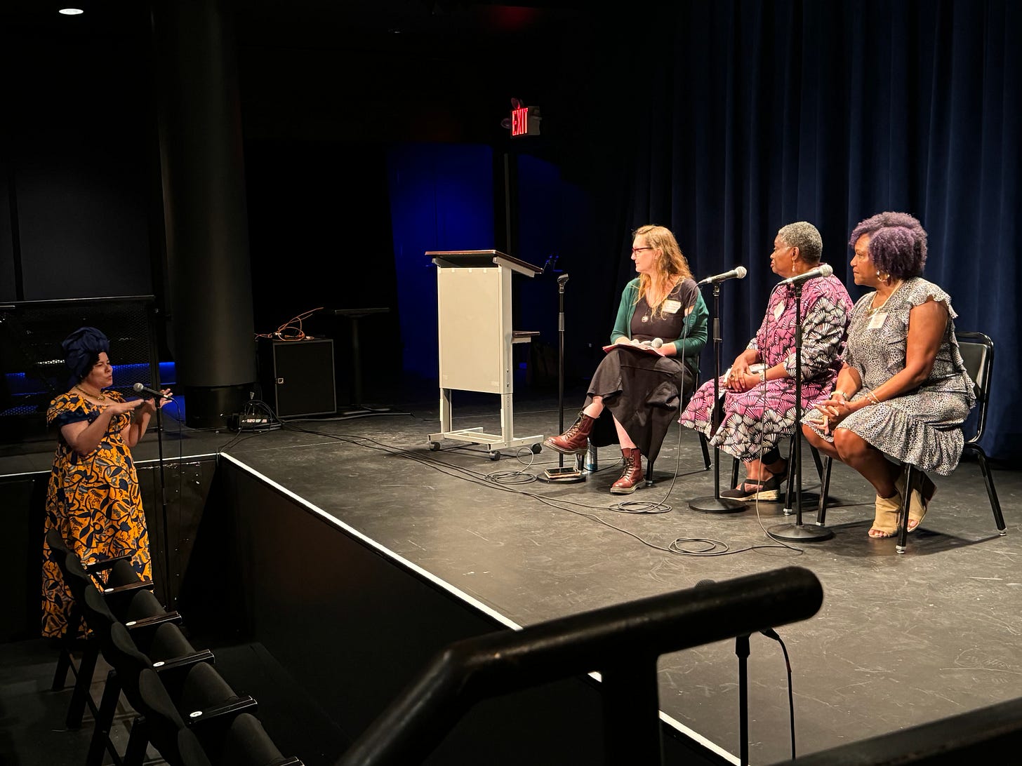 Three panelists - Executive Producer Tia A. Smith, JASNA's Renata Dennis, and the Austen Connection's Janet Saidi sit on a lit stage, looking out into the audience of a theater space, where a woman is gesturing and asking the panel a question. Three panelists - Executive Producer Tia A. Smith, JASNA's Renata Dennis, and the Austen Connection's Janet Saidi sit on a lit stage, looking out into the audience of a theater space, where a woman is gesturing and asking the panel a question.