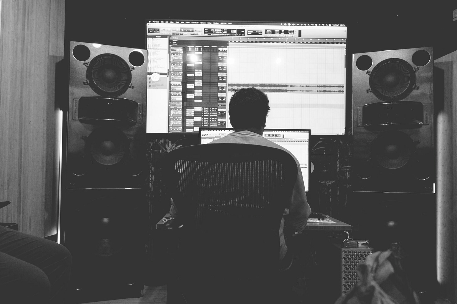 person's back is to us, sitting at table in front of computer monitor that is in front of a huge monitor tracking sounds and is flanked by large speakers. He appears to be editing music.