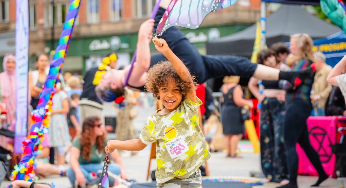 A boy wearing a brown and yellow t-shirt holding a butterfly wing fabric in the sky, with a crowd of people behind him A boy wearing a brown and yellow t-shirt holding a butterfly wing fabric in the sky, with a crowd of people behind him