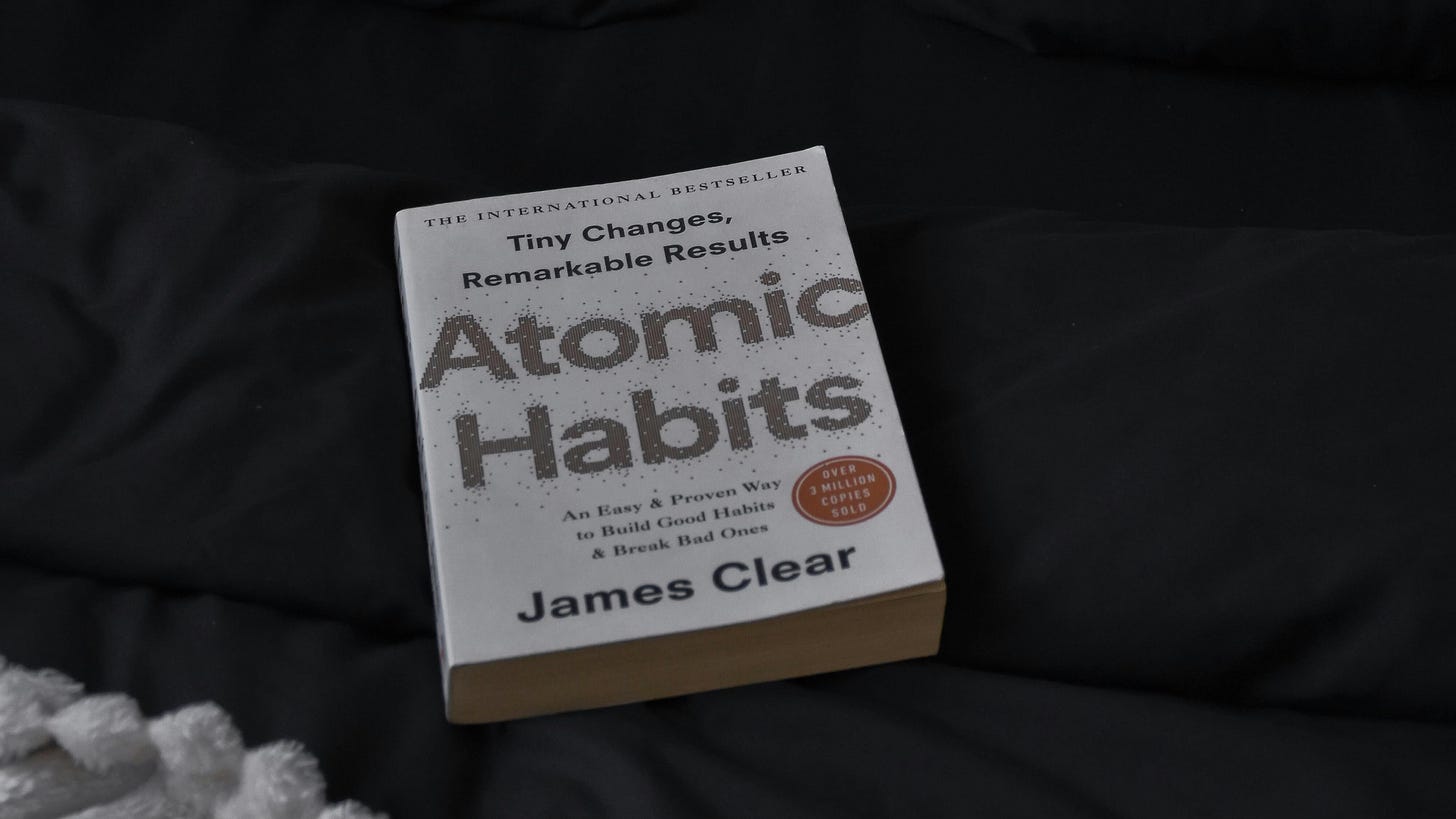 A close-up, top-down shot of the book "Atomic Habits" by James Clear resting on dark, textured fabric with a white knit blanket visible in the corner.