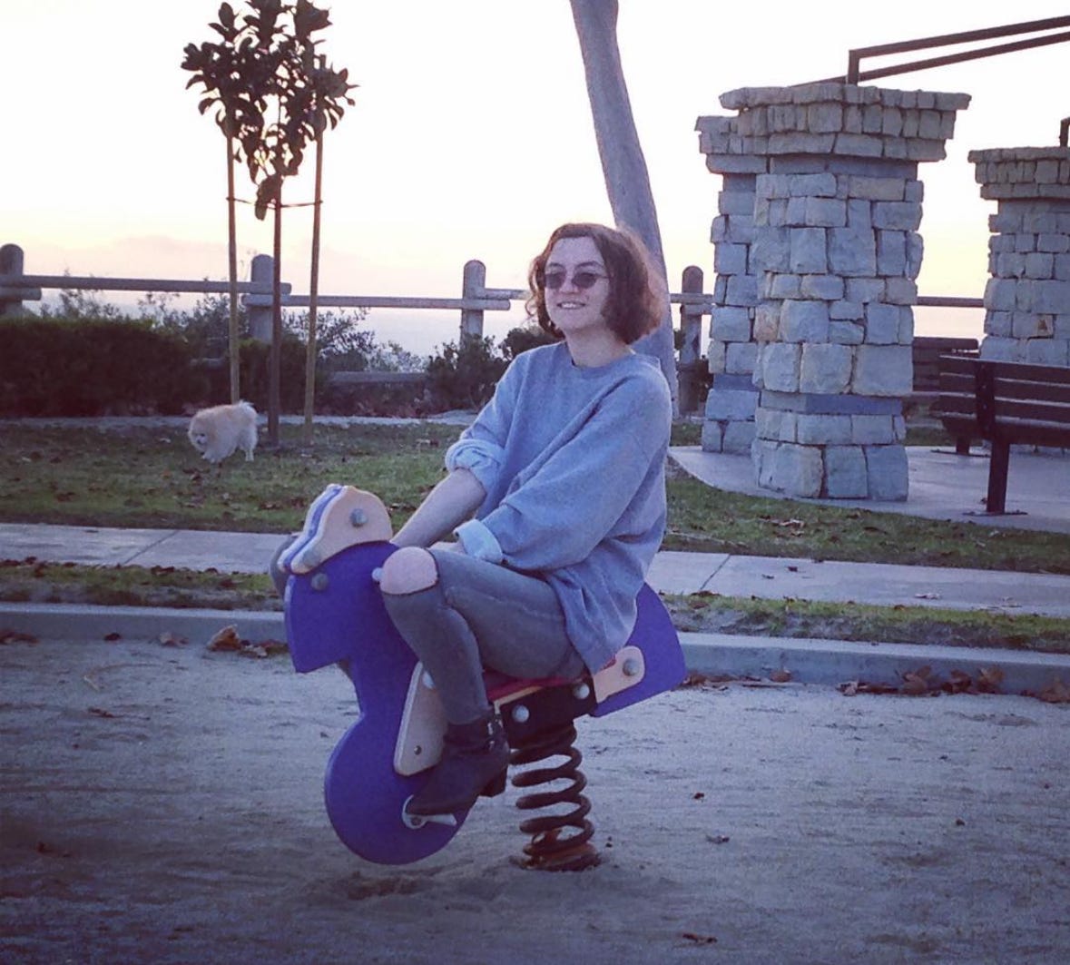 Young woman sitting in a park on a child's bouncy toy thing. Wearing a grey sweatshirt, grey pants with a hole at the knee, and ankle booties. Young woman sitting in a park on a child's bouncy toy thing. Wearing a grey sweatshirt, grey pants with a hole at the knee, and ankle booties.