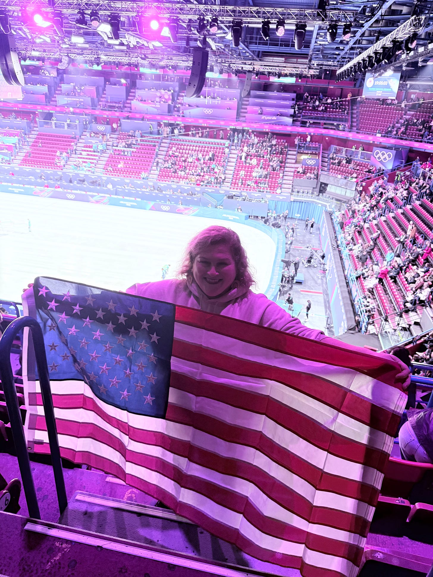 Jamie holding an American flag in an arena with an ice rink and the Olympic rings behind her