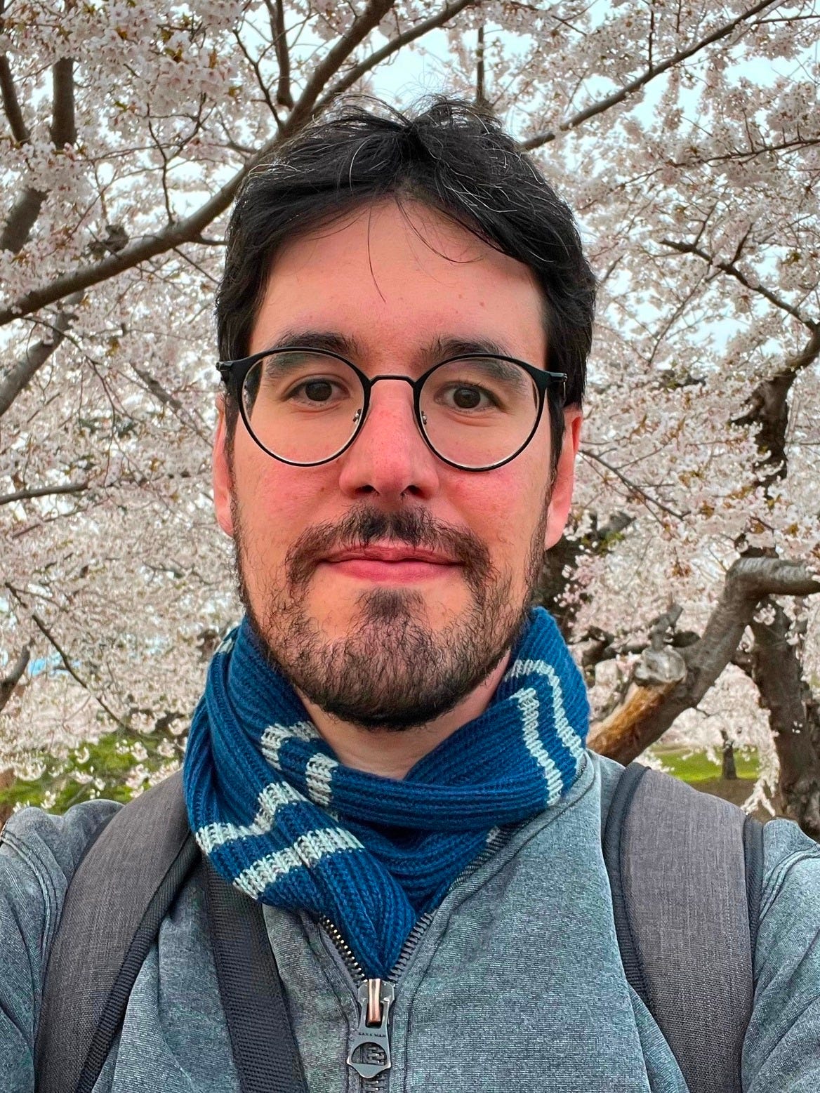 Murillo Andrade - Descrição da Imagem: Homem branco, cabelo castanho escuro e curto, óculo preto e redondo. Está vestindo uma blusa cinza e um cachecol azul e branco. Ao fundo cerejeiras brancas. Murillo Andrade - Descrição da Imagem: Homem branco, cabelo castanho escuro e curto, óculo preto e redondo. Está vestindo uma blusa cinza e um cachecol azul e branco. Ao fundo cerejeiras brancas.