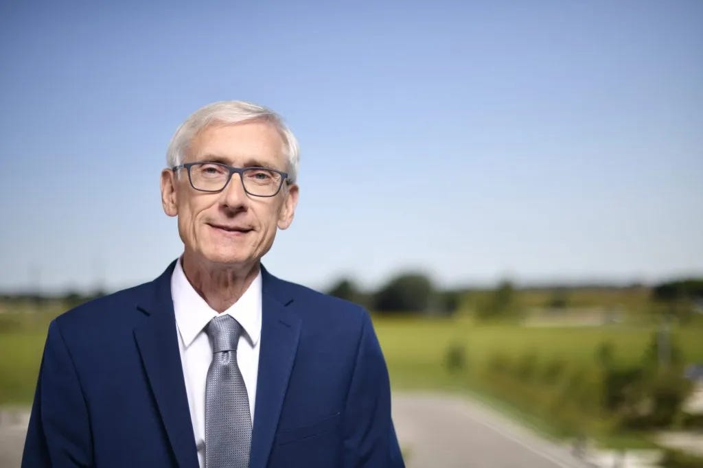 Wisconsin Governor Tony Evers stands outdoors in a navy suit and grey tie, with a green landscape and blue sky behind him.