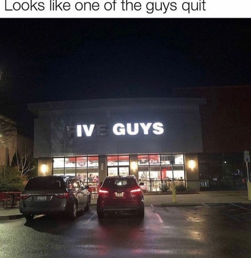 A nighttime photo of a Five Guys restaurant exterior. The building has a sign with the text "IV E GUYS" illuminated above the entrance. Cars are parked in front of the restaurant, and the parking lot is wet, reflecting the lights.