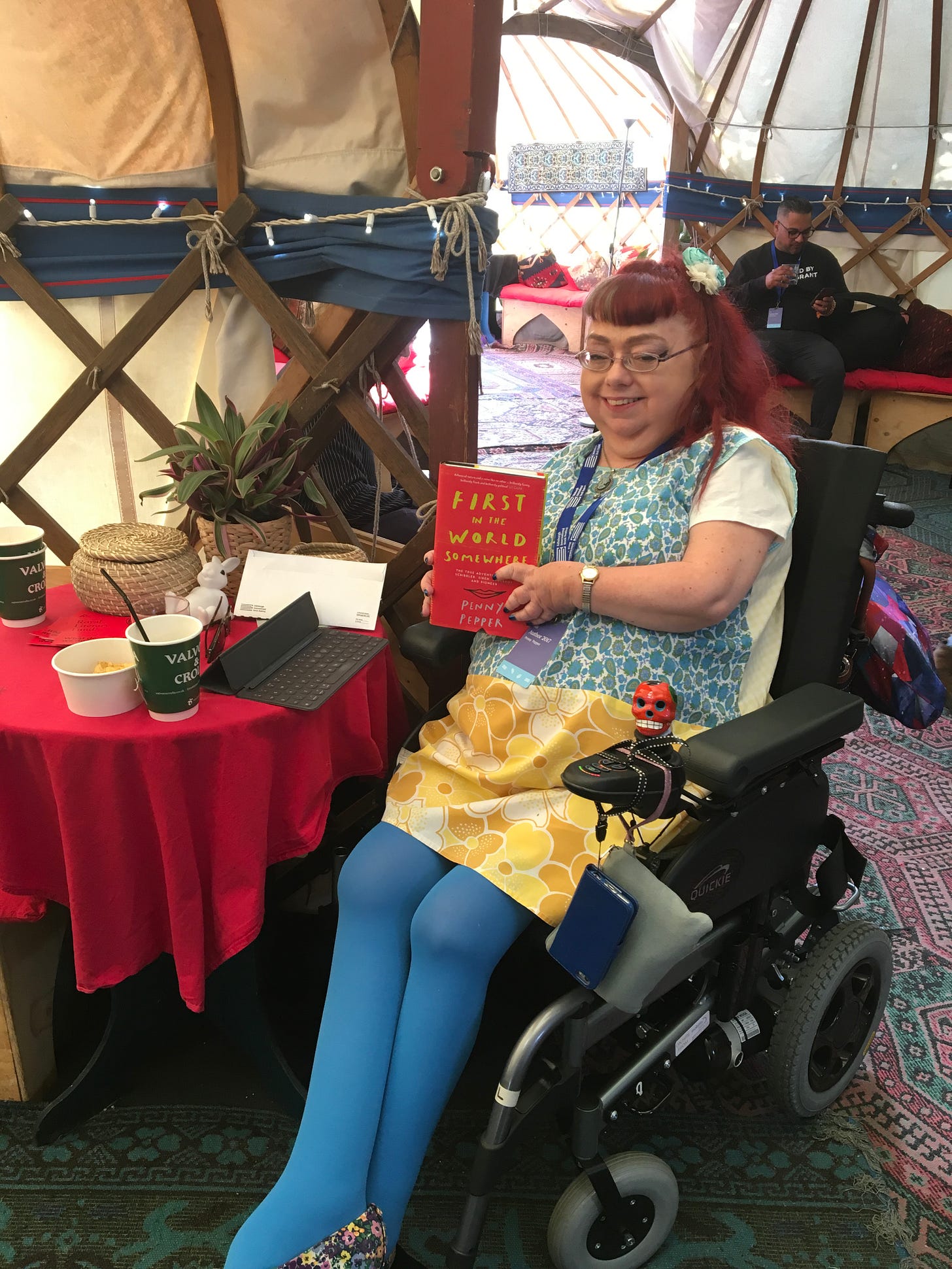 Penny sits in her wheelchair in the famous Authors’ Yurt at the Edinburgh International Book Festival. The yurt is a warm circular tent-like structure held up by wooden posts. This one is very large, and in the background there’s a fridge and facilities for refreshment on a table covered with a white tablecloth with large multi-coloured polkadots. There are carpets, cushions and throws. Penny wears blue tights and a vintage sleeveless shift dress in yellow and blue. Her hair is in a ponytail with a flower. She holds up a copy of her book, First in the World Somewhere.Penny sits in her wheelchair in the famous Authors’ Yurt at the Edinburgh International Book Festival. The yurt is a warm circular tent-like structure held up by wooden posts. This one is very large, and in the background there’s a fridge and facilities for refreshment on a table covered with a white tablecloth with large multi-coloured polkadots. There are carpets, cushions and throws. Penny wears blue tights and a vintage sleeveless shift dress in yellow and blue. Her hair is in a ponytail with a flower. She holds up a copy of her book, First in the World Somewhere.