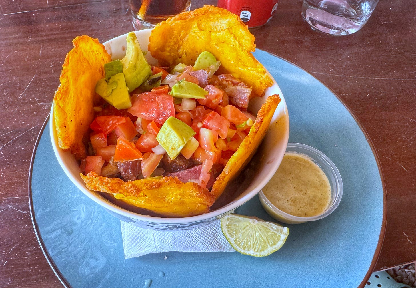 A bowl containing a mix of rice, beans, pork, avocado, and pico de gallo surrounded by fried plantains and a wedge of lime