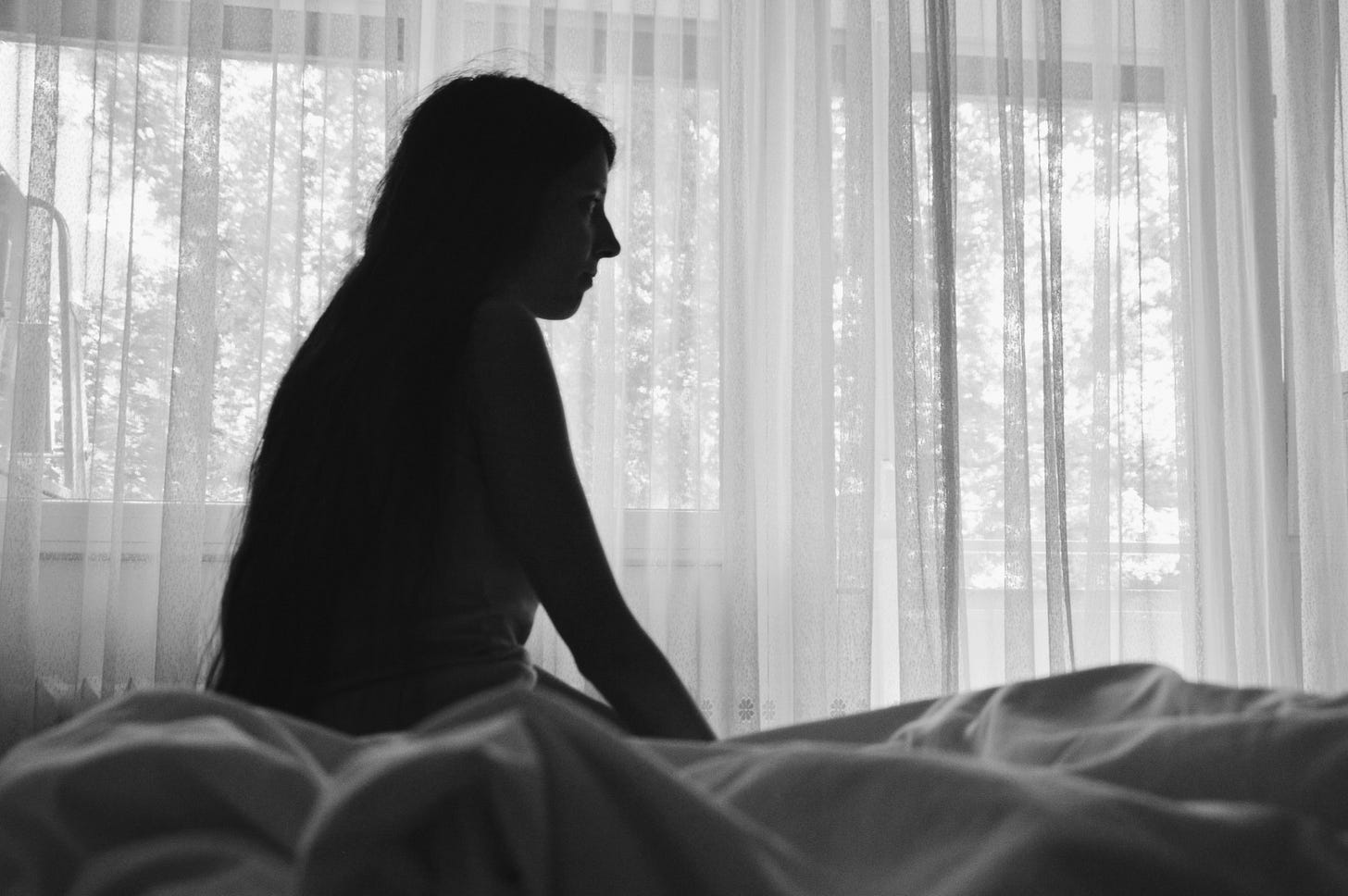 A woman sits in shadow on a bed. She is alone in a white room. A woman sits in shadow on a bed. She is alone in a white room.