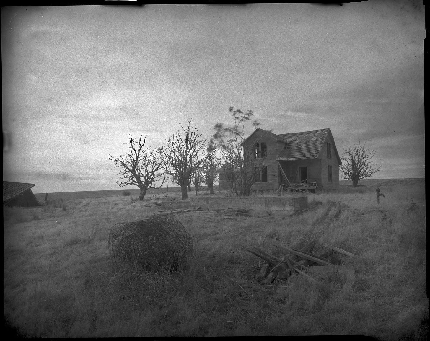 Old looking photo of an abandoned farmhouse. A few bare trees and a bail of barbed wire are in the foreground.