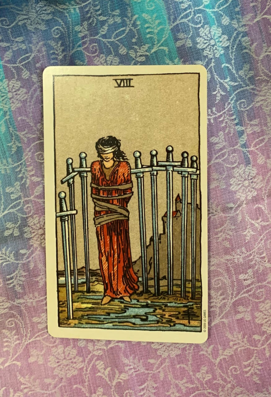 a white woman in a red dress and a blindfold is surrounded by 8 swords she’s standing on a scrap of sand amid water. The card rests on a rainbow silk scarf.