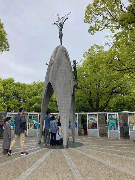 Photo of the Children's Peace Memorial, a tall concrete structure with a crane on top. Colorful strings of folded cranes. Museum display with tiny cranes foleded by Sasaki.