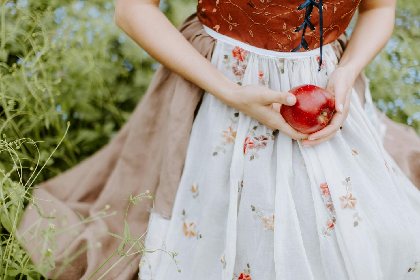A young woman shown from mid-torso down kneeling an the grass. She is wearing a red corset top with golden florals, a brown linen skirt, and a white apron with florals on it. In her hands she holds an apple.
