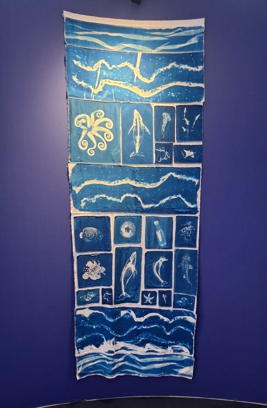 Foto. Mostra um tecido com ilustrações em branco e fundo azul (ciano). Há algumas baleias, estrela do mar, peixes recifais, polvos, entre outros