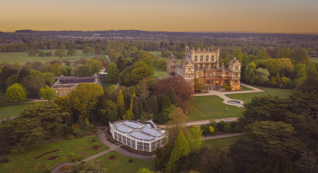 A wide shot of Wollaton Hall with trees around it and a sunset sky above it
