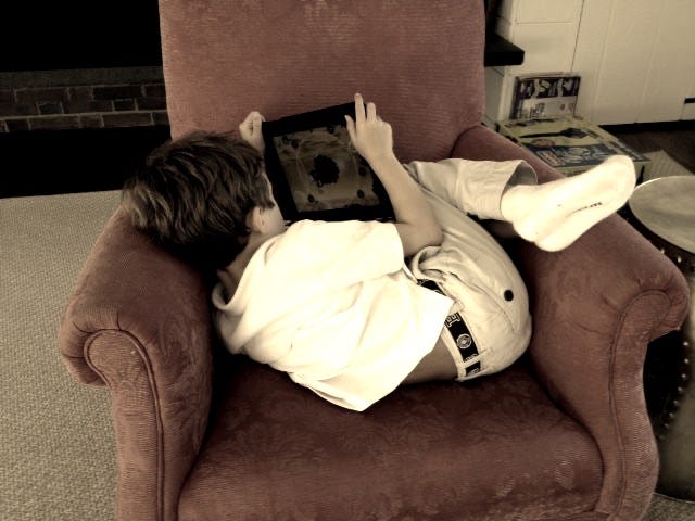 This may contain: a young boy laying on top of a red chair holding an electronic device in his hands This may contain: a young boy laying on top of a red chair holding an electronic device in his hands