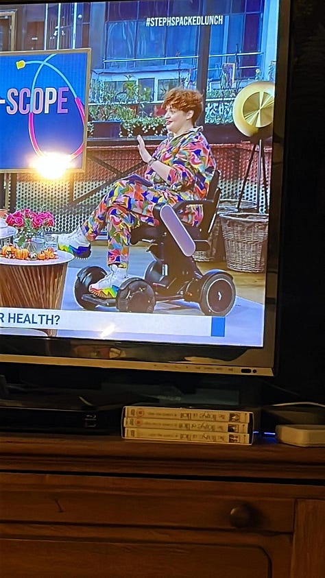 1) Hannah sits in a powerchair with pink sides, wearing a cord rainbow boilersuit and rainbow converse, on the TV on the Steph’s Packed Lunch Set. 2) Hannah dressed as Enid Sinclair, with pink wig and false rainbow nails 3) Hannah stands by her chair in a disabled loo, taking a photo in the mirror, wearing a sparkly green sleeveless v neck top and a big puffy green net skirt