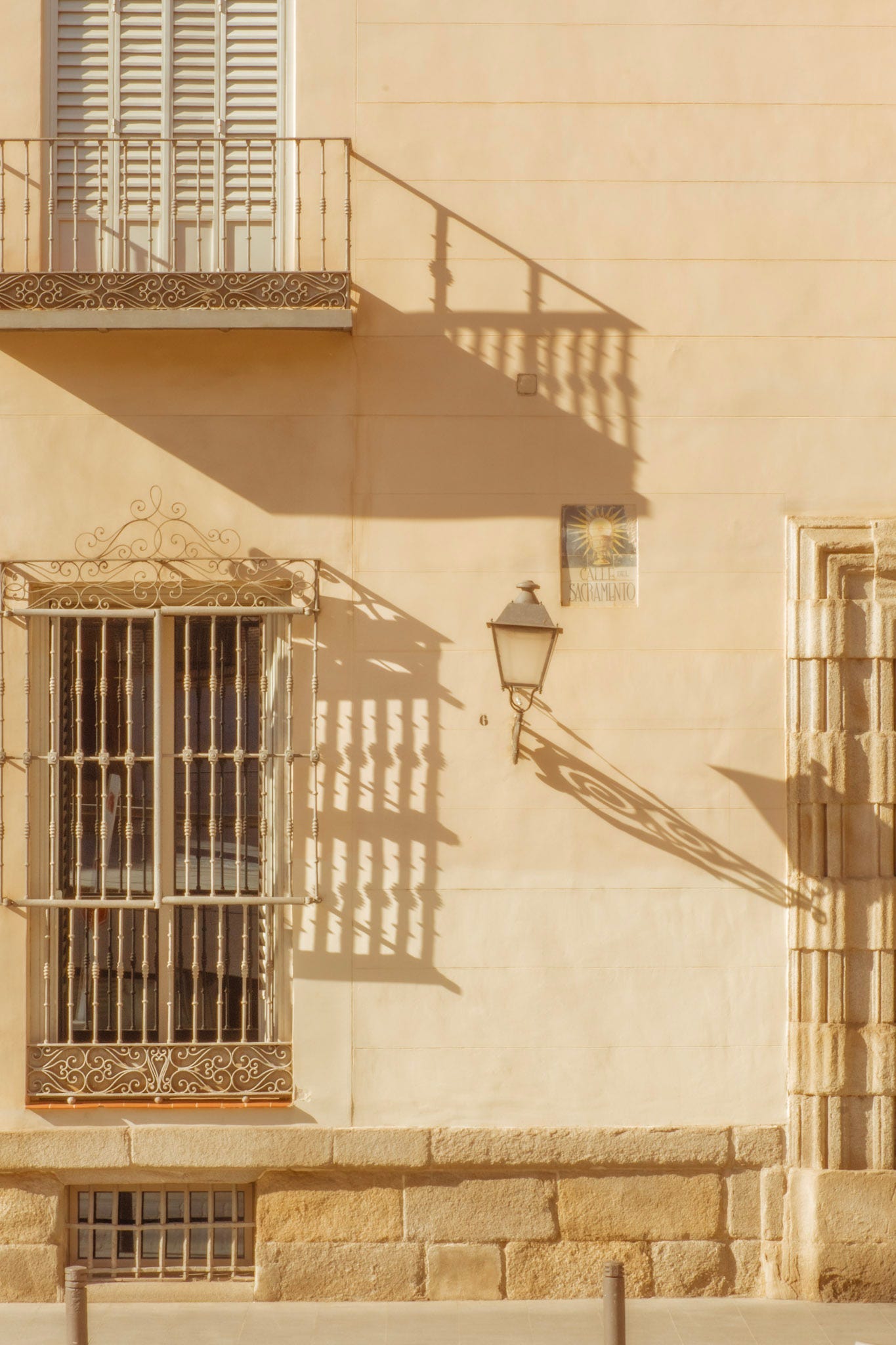 Sunlit historic building façade in Spain with wrought-iron balconies, shuttered windows, and a decorative lantern casting long afternoon shadows across a cream-coloured wall.