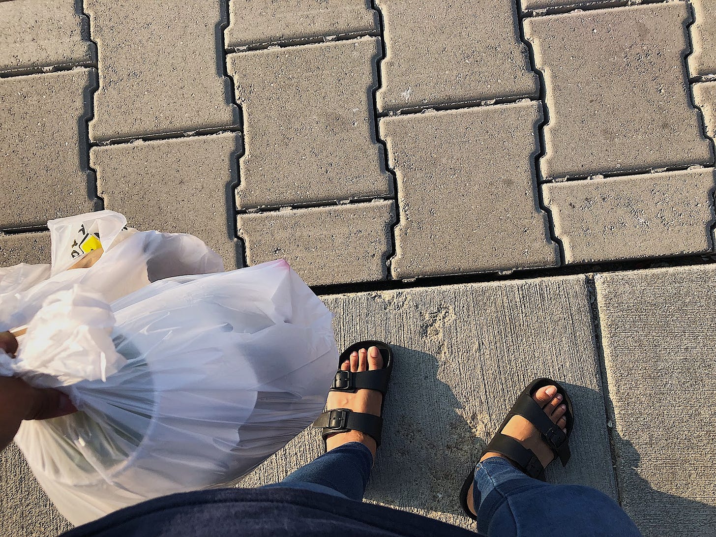 A photo of pavement, feet in sandals, and a bag of sushi to go A photo of pavement, feet in sandals, and a bag of sushi to go