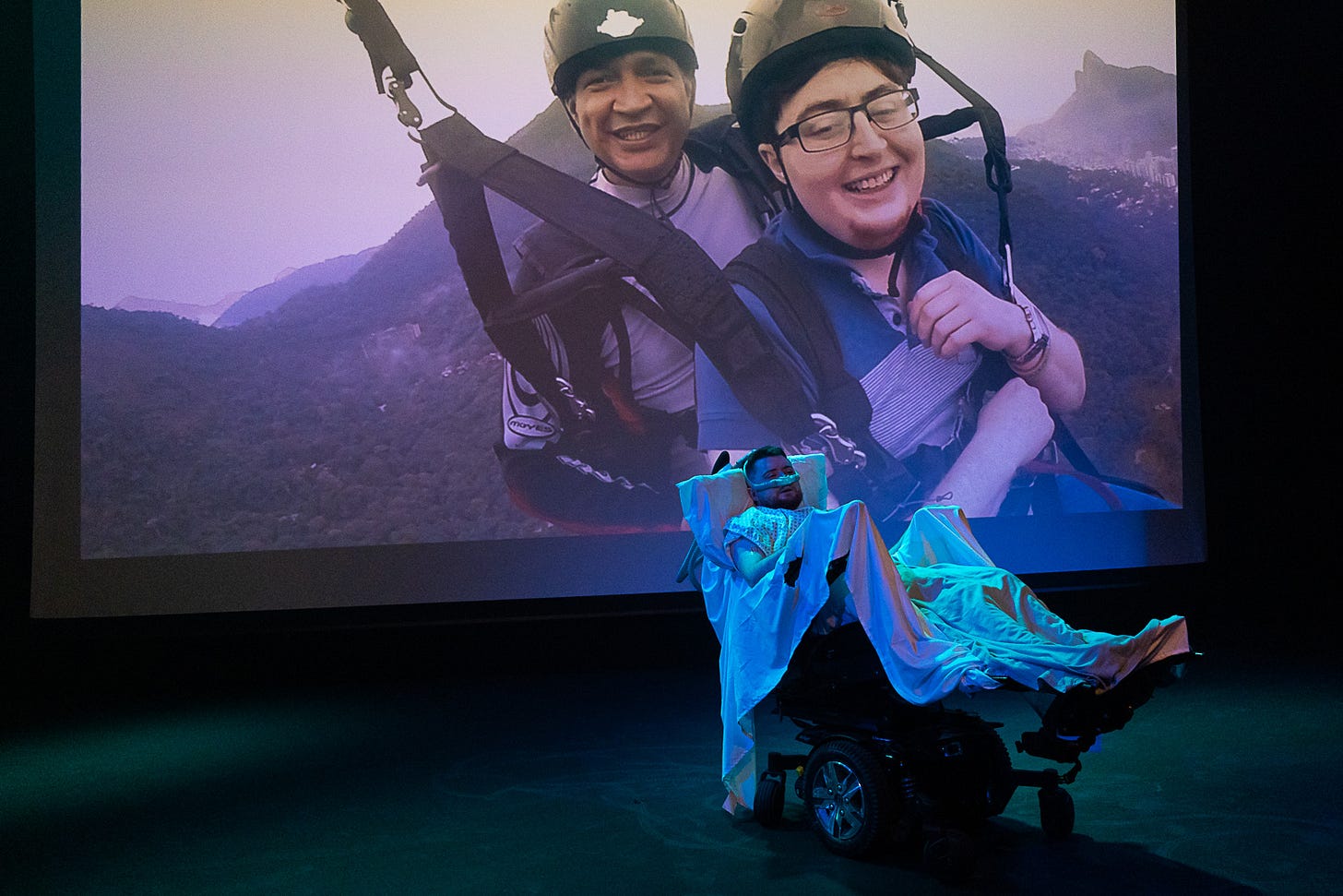 Projected onto a screen, Jamie Hale with a strange man paragliding through mountains. In front of that, Jamie in their electric wheelchair, set up like a hospital bed.